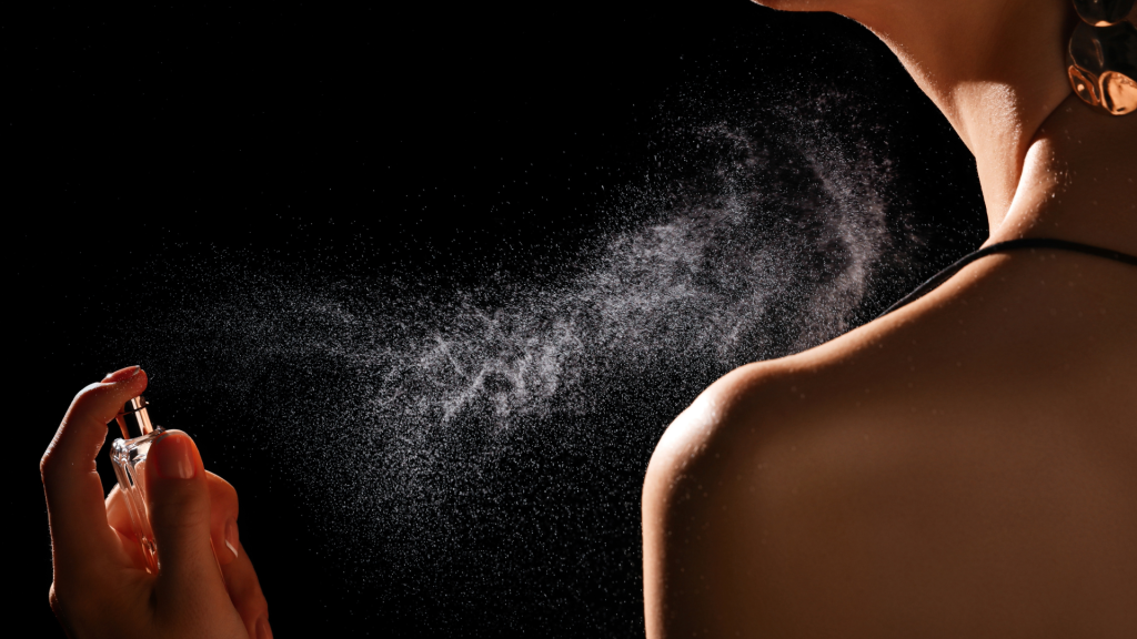 Cropped side view of a woman misting cologne on her neck and shoulder against a black background, with the perfume mist highlighted by lighting.