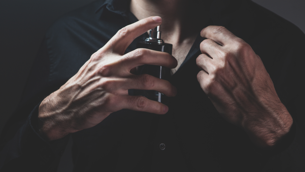 Close-up of a man in a black shirt spraying cologne on his chest, with focus on his hands holding the bottle in low, moody lighting.